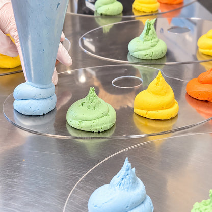 Blue buttercream frosting being squeezed onto plastic mixing pallets where green, yellow, and orange frosting mounds already exist.