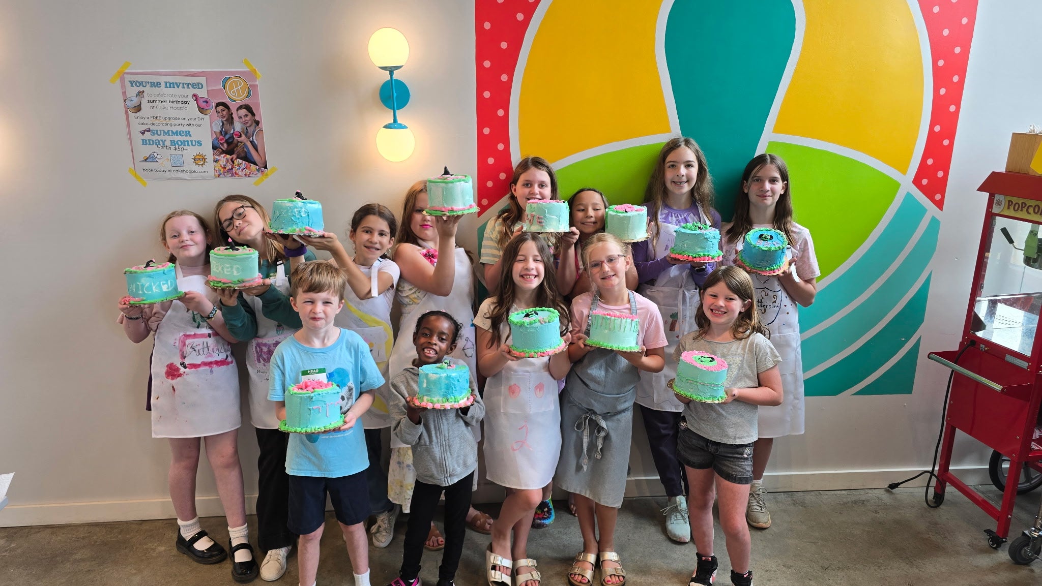 A group of elementary age kids wearing white aprons proudly holding decorated cakes in a colorful indoor setting with a large yellow, green, and red abstract mural on the wall.