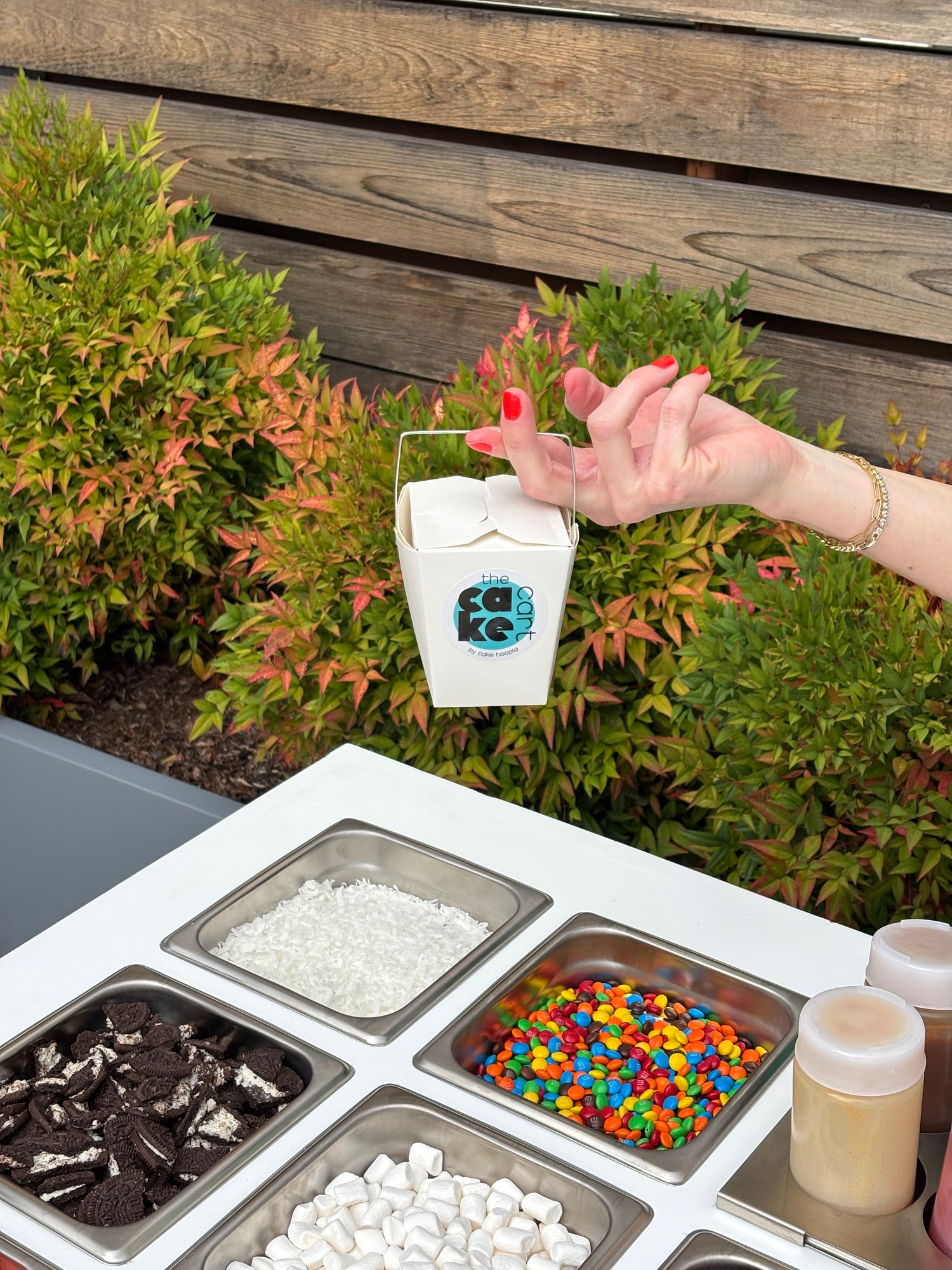 Cake Hoopla's mobile dessert cart set up for an event in the Portland area with cake, fun toppings, and yummy drizzles and a handy to-go box held above the toppings.