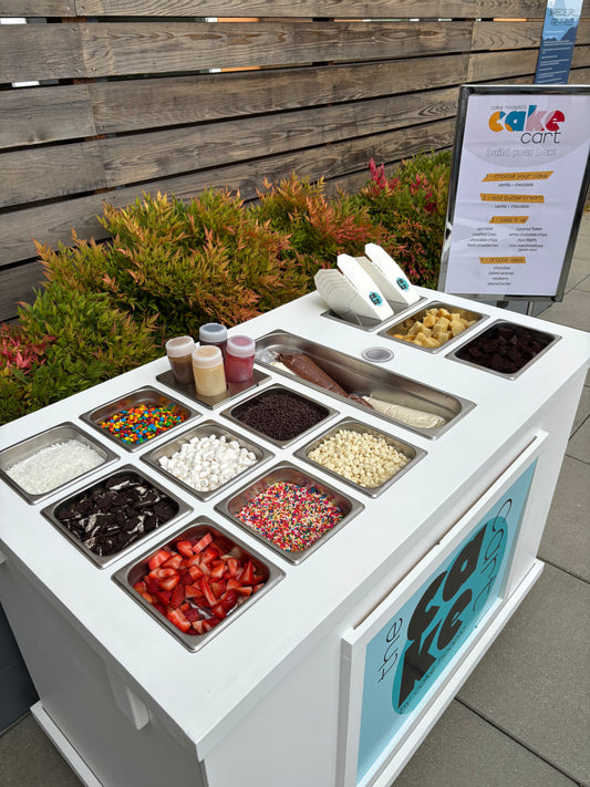 Cake Hoopla's mobile dessert cart set up for an outdoor event in the Portland area with cake, fun toppings, yummy drizzles, and a standing menu sign.