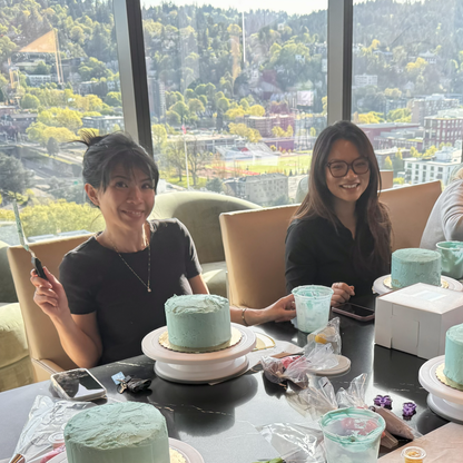 Two women decorating blue cakes with the skyline of Portland in the background.