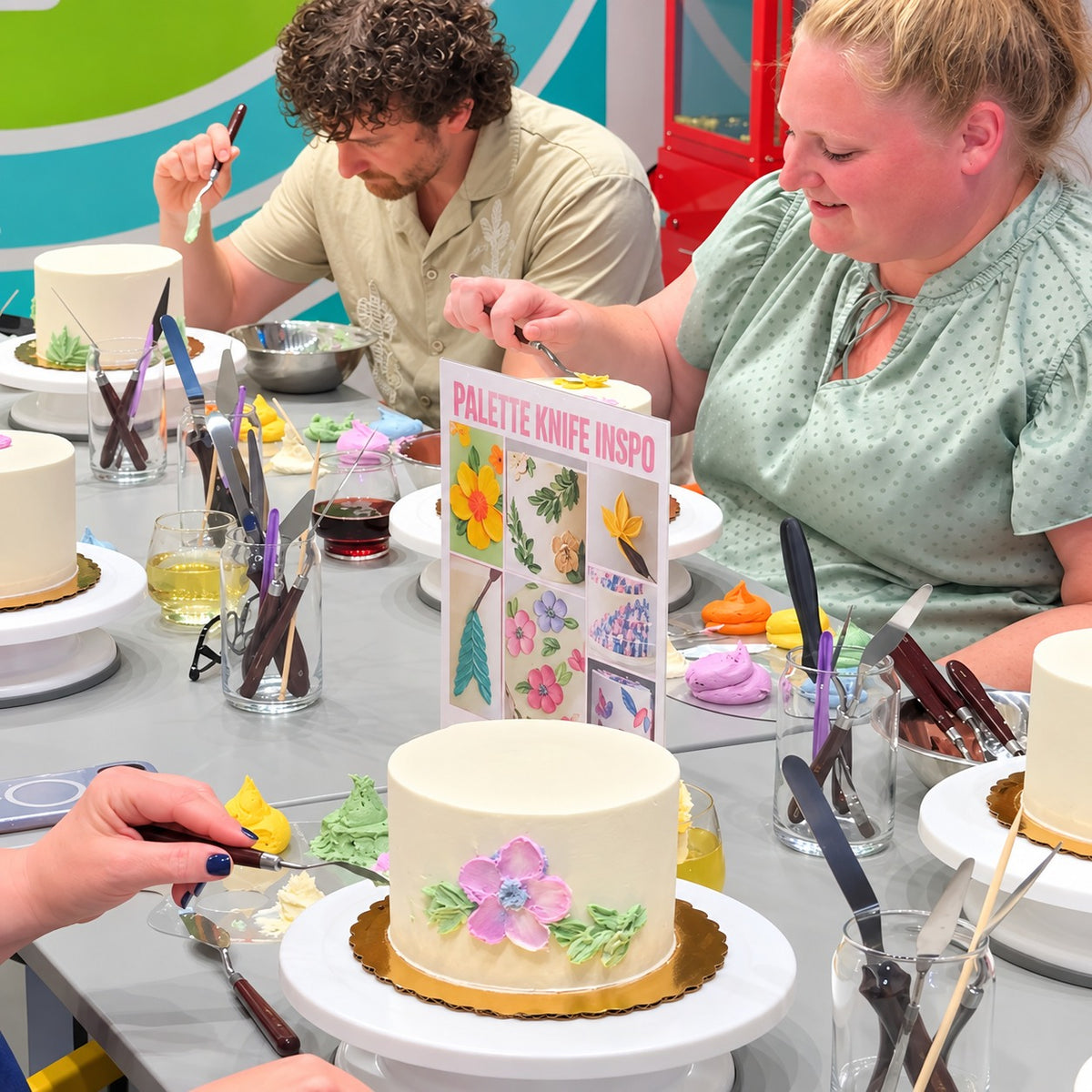 A table contains 5 round white cakes on turntables and a man and a woman are pictured applying buttercream paint with pallet knives to their cakes in a studio setting and wine glasses and tools atop the table.