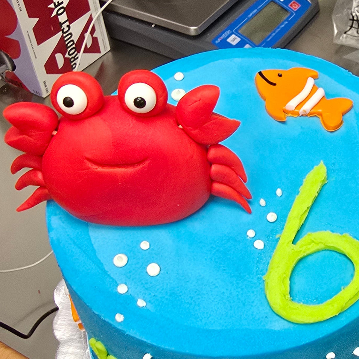 A cake decorated with red fondant shaped into a crab, yellow fondant shaped into a fish, and green fondant icing to represent seaweed, part of a cake decorating workshop.