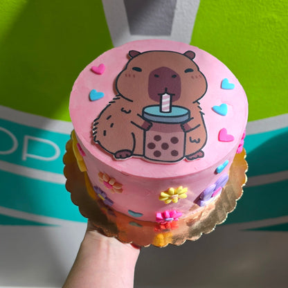 A hand holding a pink frosted cake decorated with a cartoon capybara holding a bubble tea, set against a bright green and white background.