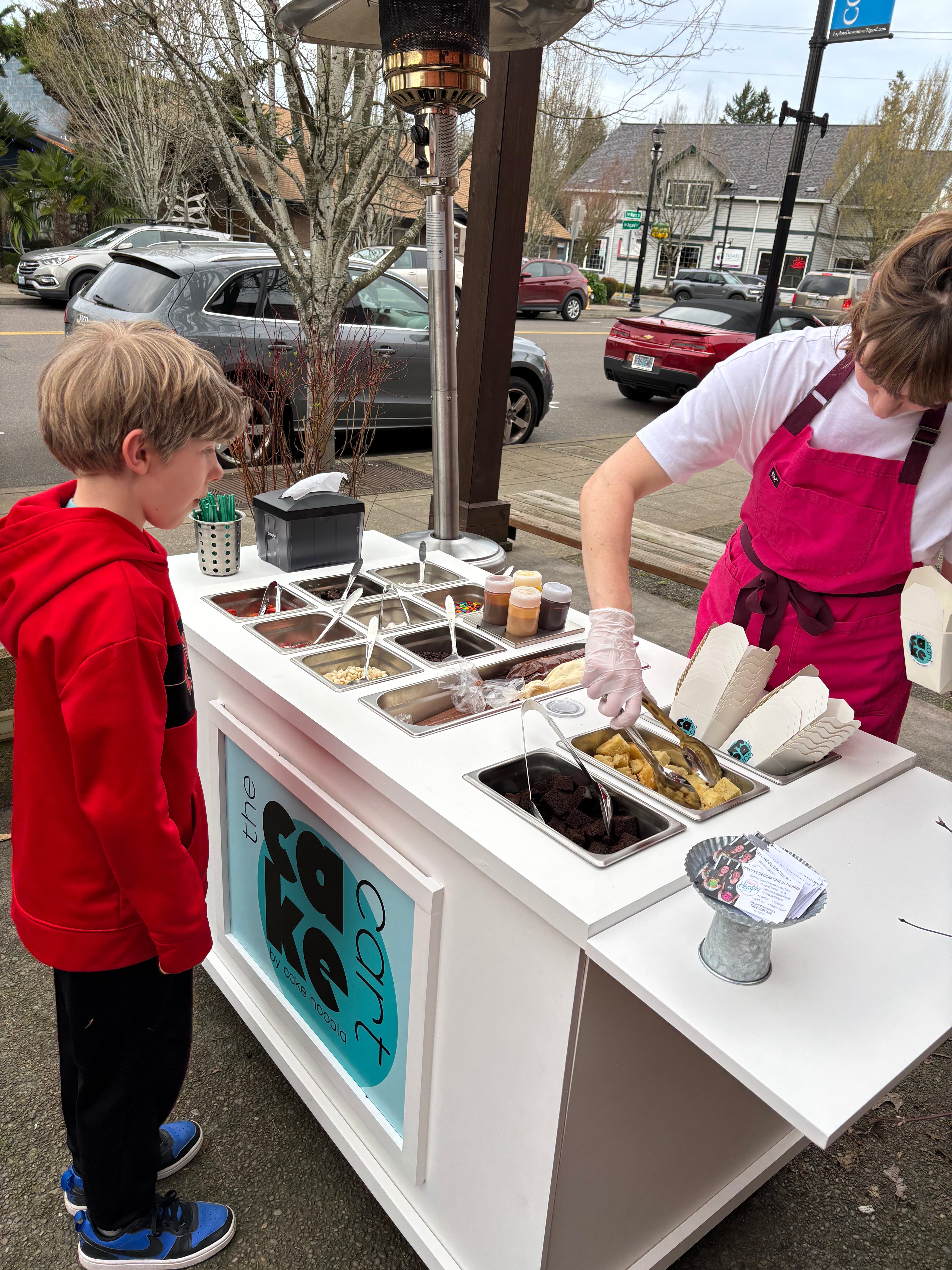 Cake Hoopla's mobile dessert cart set up for an outdoor event in the Portland area with cake, fun toppings, and yummy drizzles, as helper serves guest.
