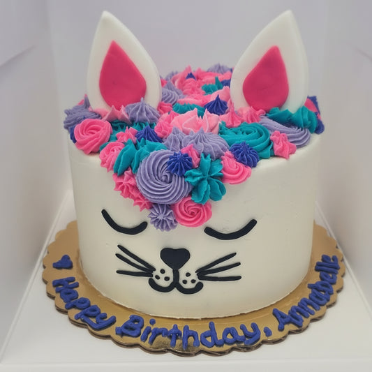 A custom cat-themed birthday cake decorated with white frosting, colorful piped flowers in pink, blue, and purple, and a cat face. The cake is topped with white frosting ears with pink linings and sits on a gold cake board with a message written on the cake board.