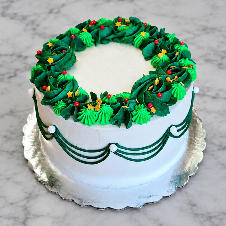 White cake with dark green drop strings, light and dark green frosting wreath, and Christmas sprinkles on a silver cake board.