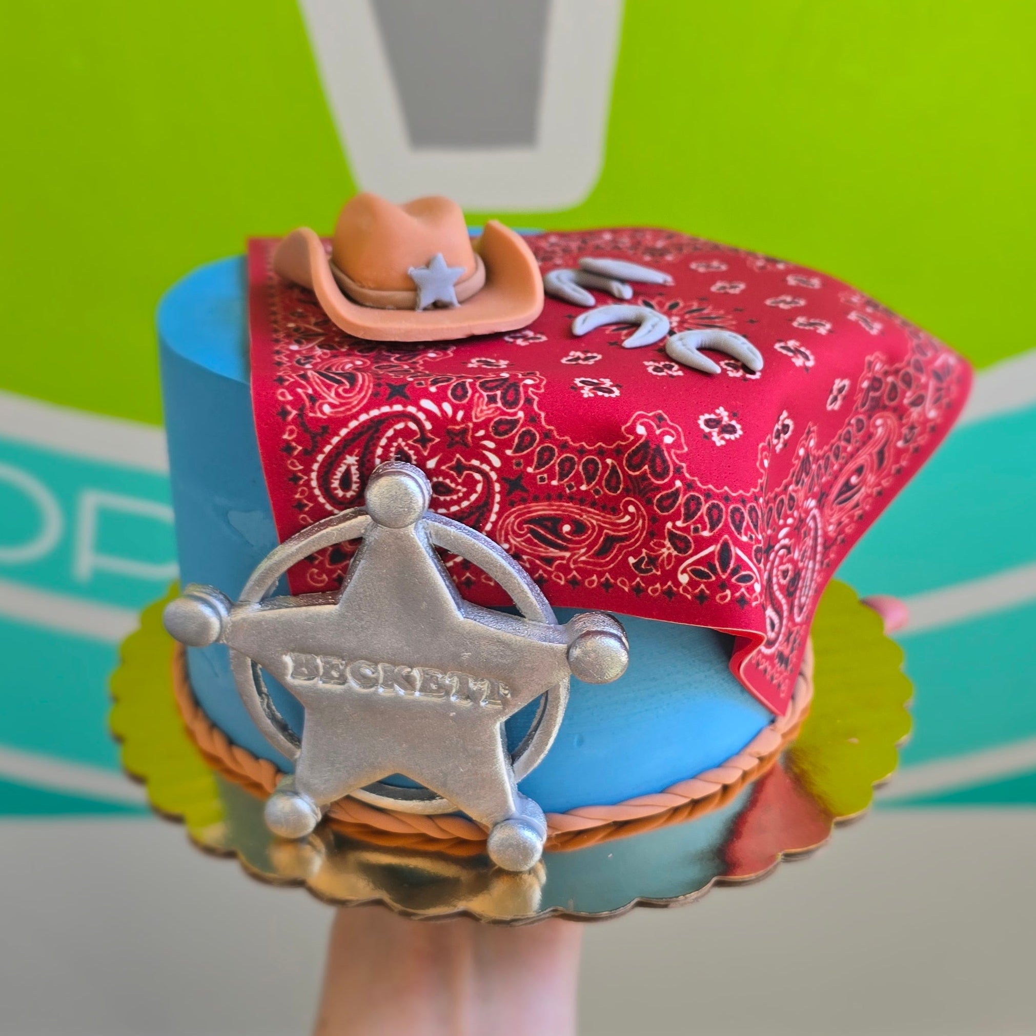 Hand holding a cowboy themed cake with a red bandana, sheriff's badge, and fondant cowboy hat with rope border around the bottom of the blue cake.