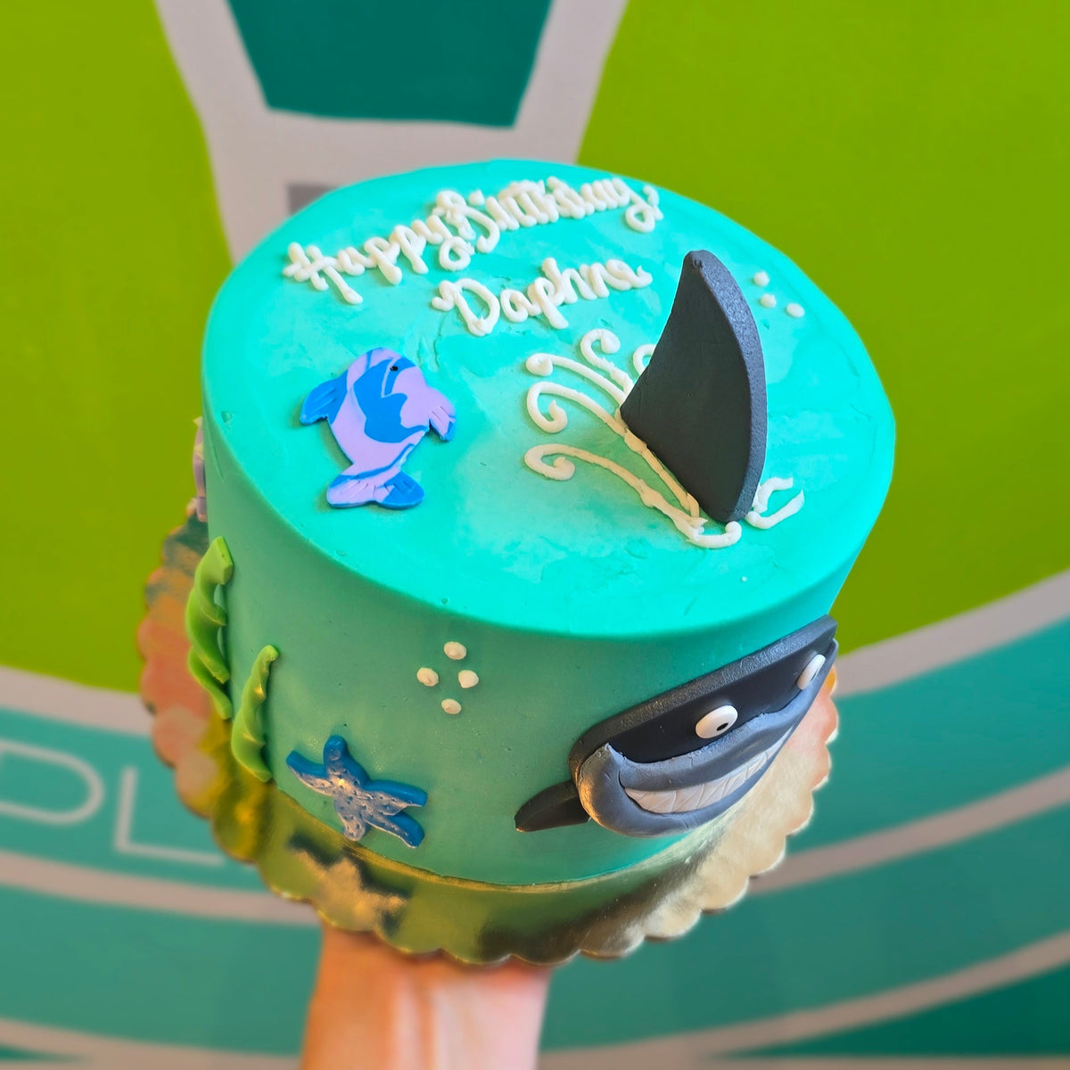 A custom underwater-themed birthday cake held up by a hand. The cake is decorated with turquoise icing, featuring a blue fish, a black shark fin, a blue-and-black shark face, and 'Happy Birthday Daphne' written in white script on top.
