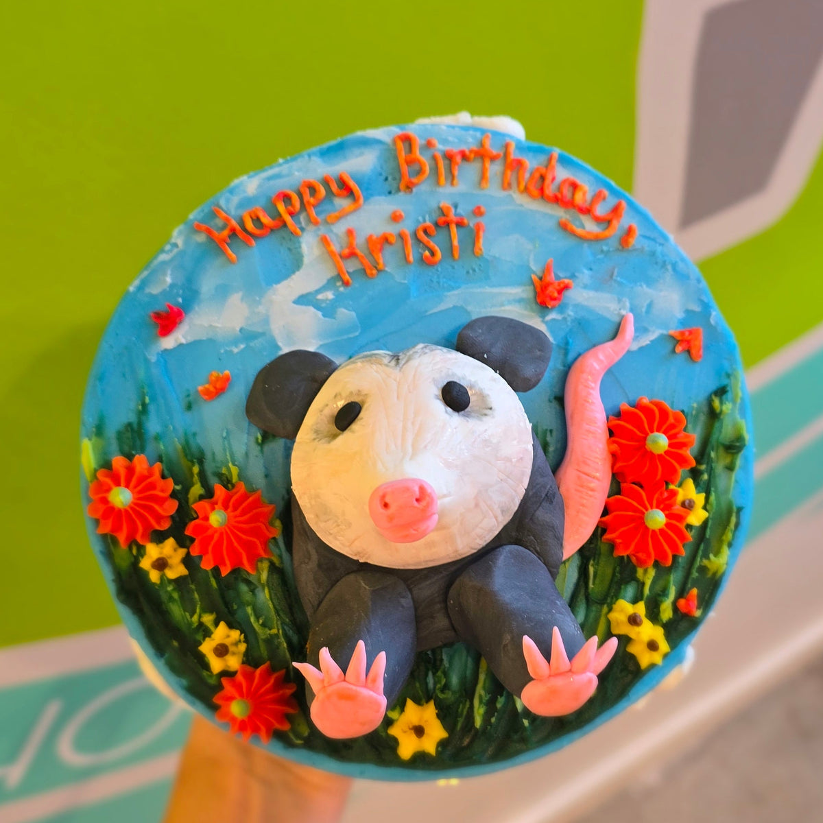 Top of a cake frosted blue with a grass and pumpkin motif and a fibdant possum popping out from the top of the coke. 
