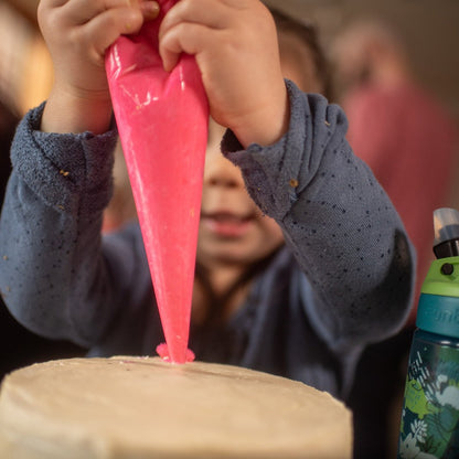 A little girl is at a custom to-go DIY cake decorating party by Cake Hoopla in the Portland area, squeezing pink frosting out of a piping bag and onto a cake. 