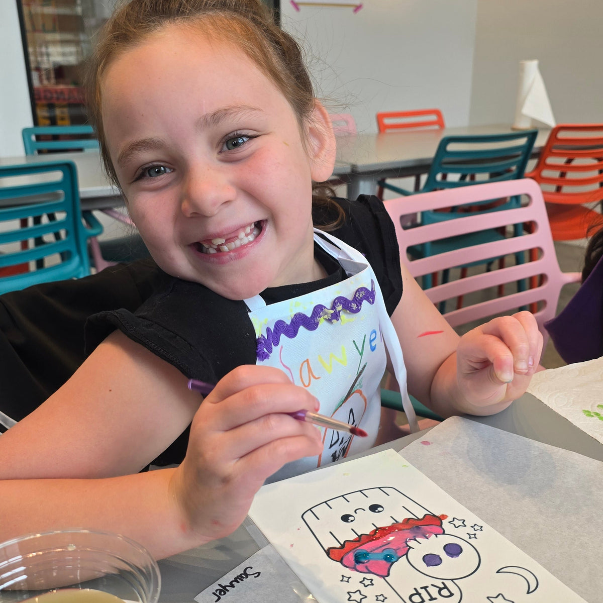 A girl smiles at the camera while holding a paintbrush in front of an edible image of spooky cupcakes that she is painting with edible pink, blue, and red paint.