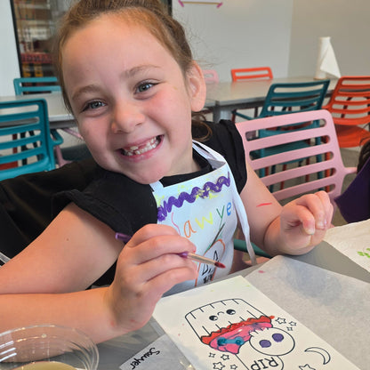 A girl smiles at the camera while holding a paintbrush in front of an edible image of spooky cupcakes that she is painting with edible pink, blue, and red paint.