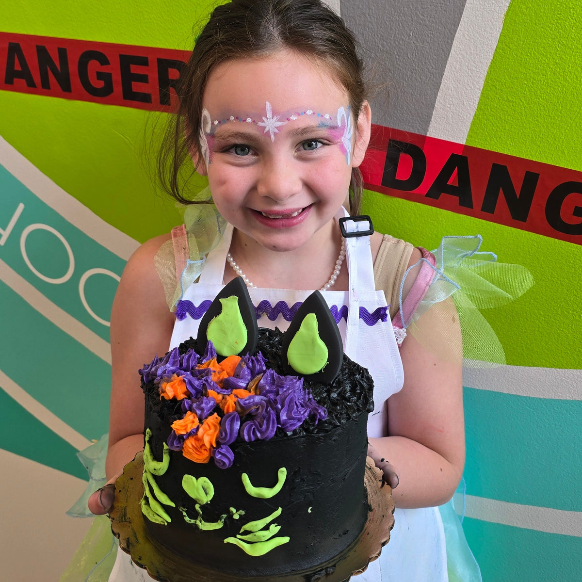 A girl dressed as a fairy holds her black cat cake made during cake camp in front of a colorful wall with "Danger" tape across it.