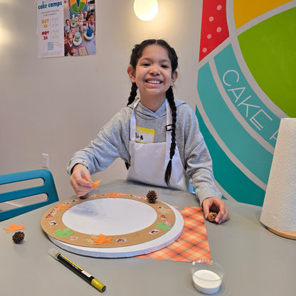 A girl smiles big at the camera as she applies pinecones and fall leaves to her harvest craft project at Cake Hoopla.