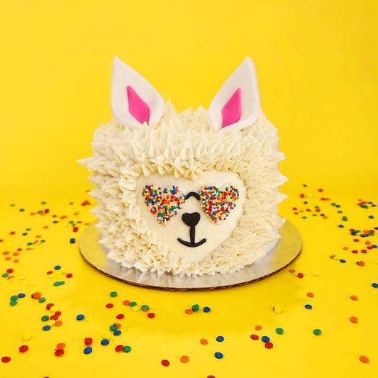 Cute llama cake with white frosting fur, heart-shaped rainbow-sprinkled sunglasses, and white ears with pink centers.