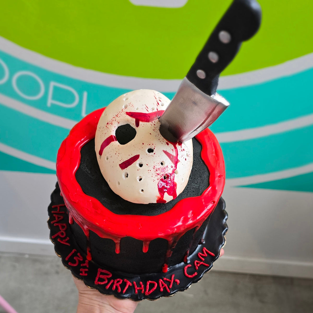 Fondant hockey mask atop a black cake with fondant butcher's knife cutting through eye socket and white chocolate blood dripping down sides.