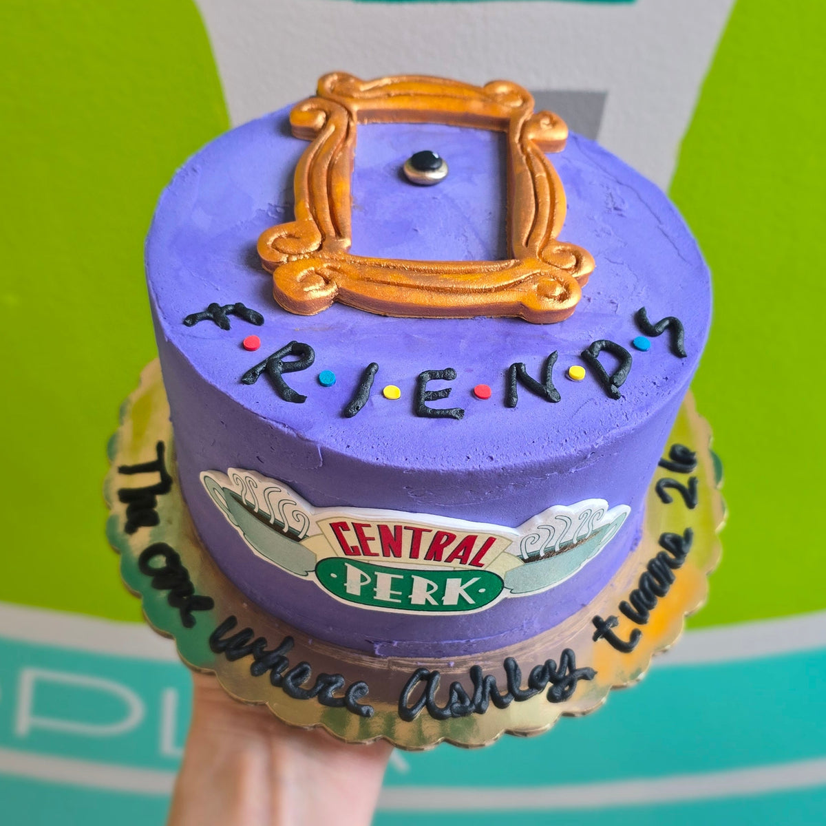 Hand holding a purple pillow with 'Friends' logo and text against a green and gray background