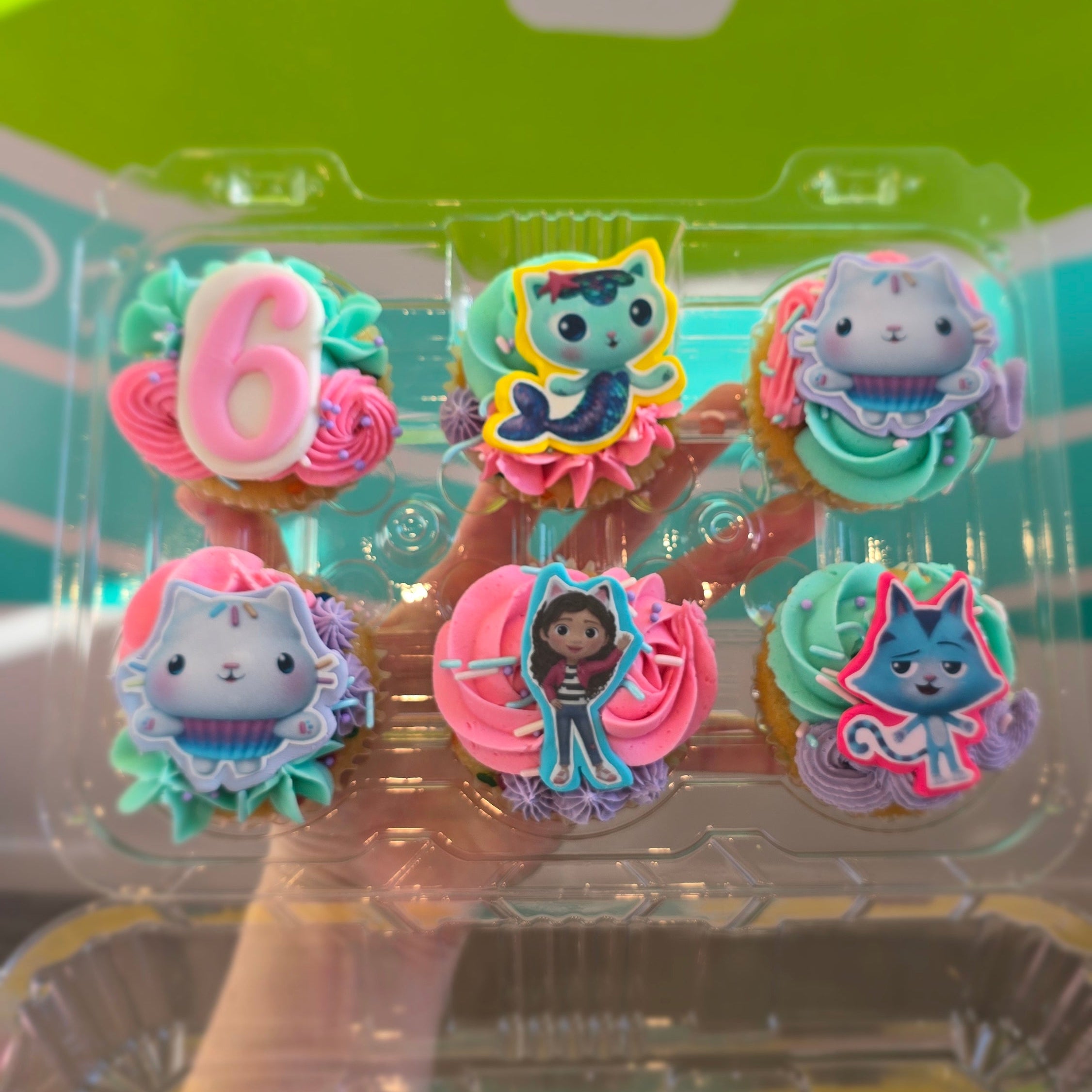Six colorful cupcakes in Gabby Dollhouse theme with characters and pink, teal, and purple frosting.