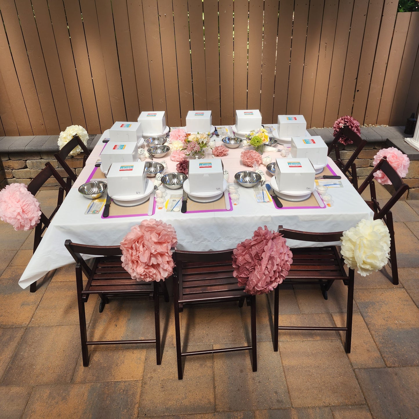 A garden-party-themed cake-decorating to-go party is set up with cakes, tools, and colorful pink  pom pom decorations.