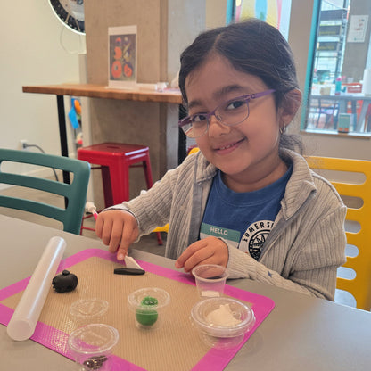 A girl smiles at the camera as she uses a fondant knife to cut black fondant on a fondant mat.
