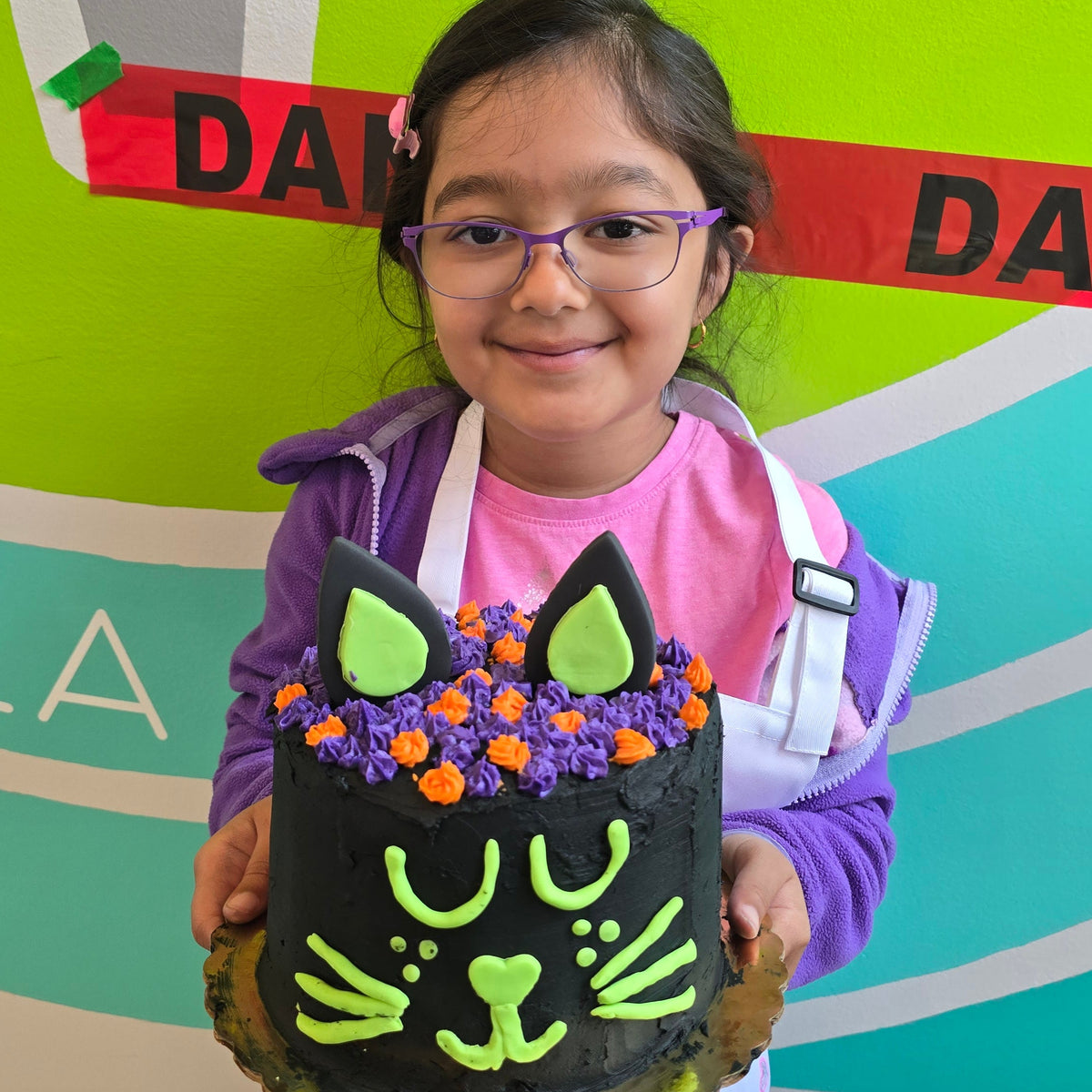 A girl stands in front of colorful background and "danger" tape while holding a black cat cake she made.