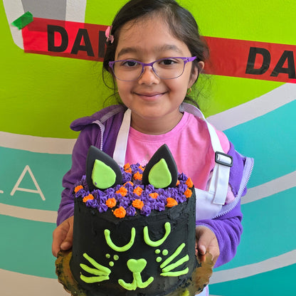 A girl stands in front of colorful background and "danger" tape while holding a black cat cake she made.