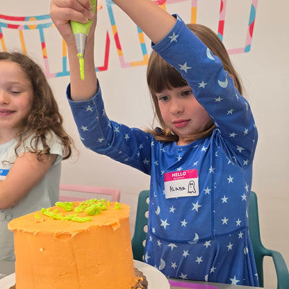 With her arms stretched high above the cake, a girl squeezes bright green frosting from a piping bag onto an orange cake.