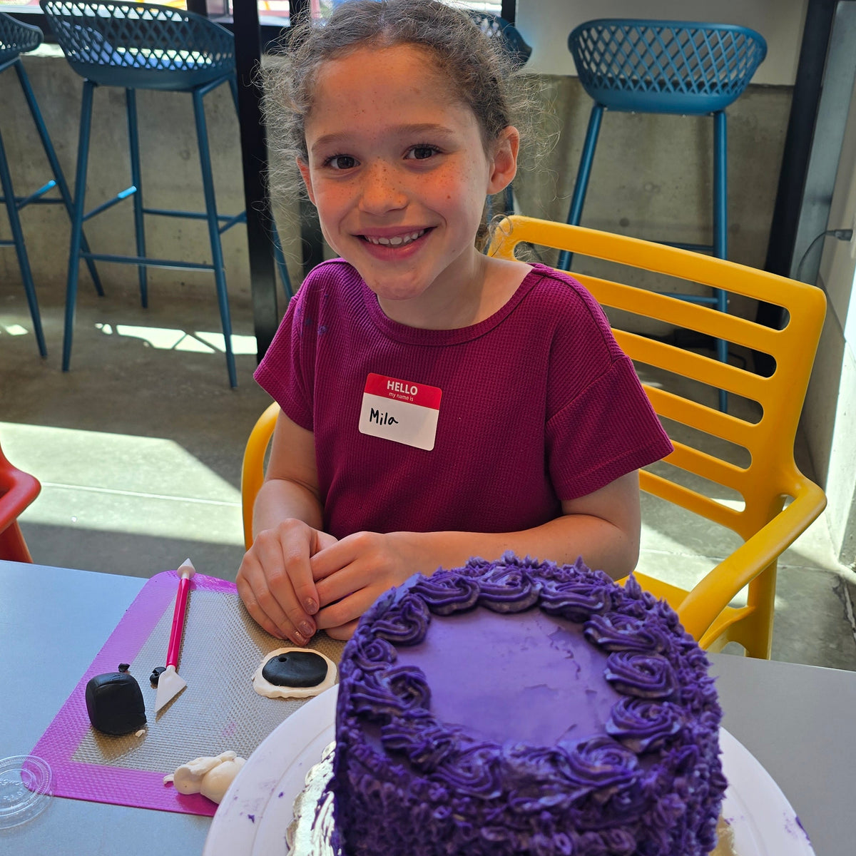 A girls miles at the camera as she shapes black and white fondant into circles for her purple-frosted cake in front of her.