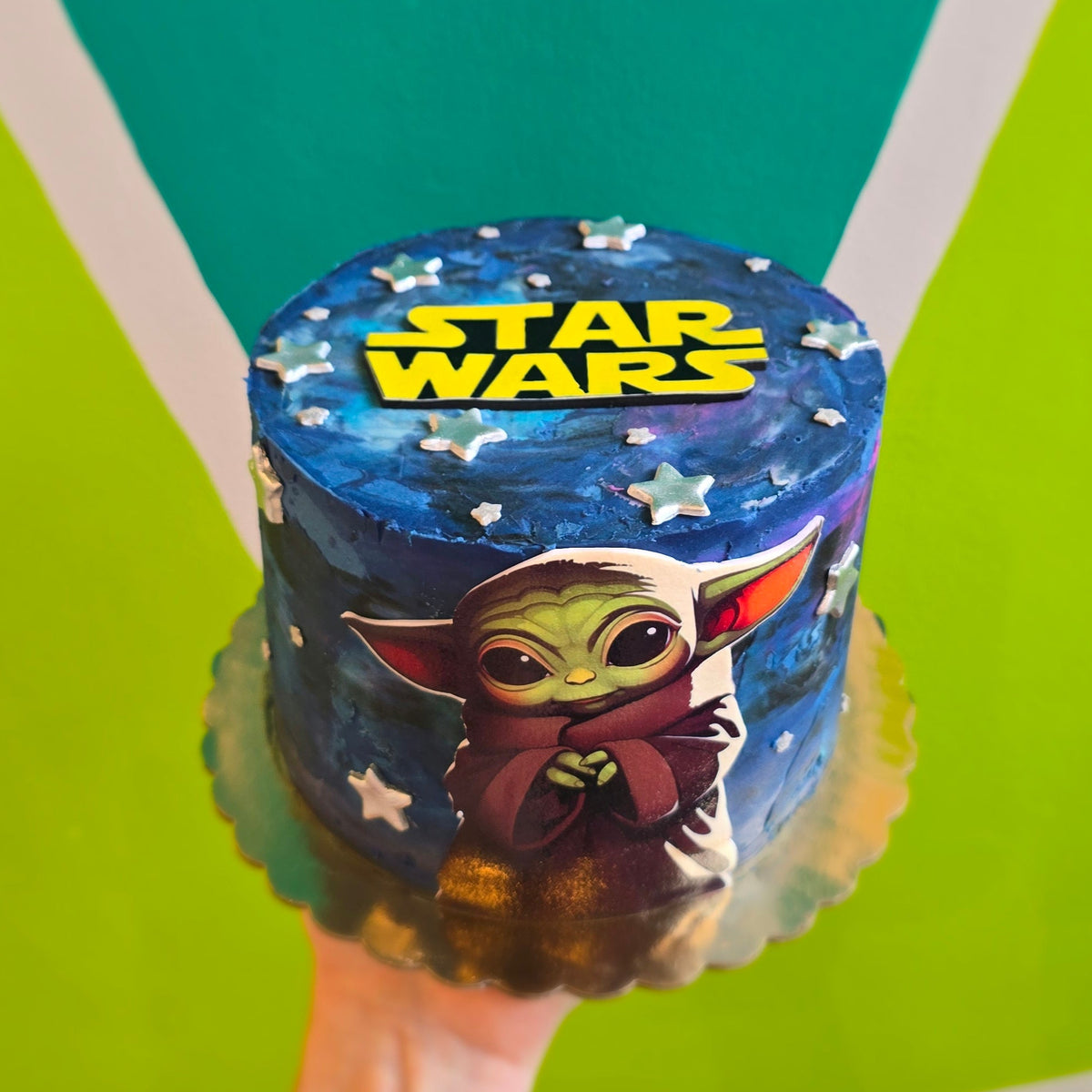 Baby Yoda (Grogu) cake with dark galactic frosting, silver fondant stars, an image of Grogu across the front and Star Wars logo on top.