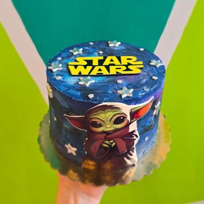 Baby Yoda (Grogu) cake with dark galactic frosting, silver fondant stars, an image of Grogu across the front and Star Wars logo on top.