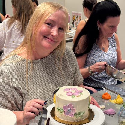 A woman smiles as the camera as she pauses applying green paint with a palette knife to a white cake in front of her on a turtable.