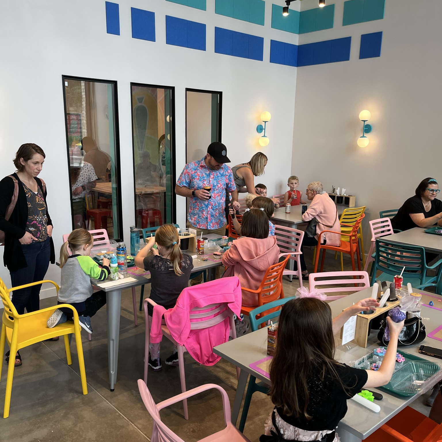 four tables are full of adults and children decorating cupcakes and having a good time. Some adults are drinking beer and wine, and kids are drinking chocolate milks