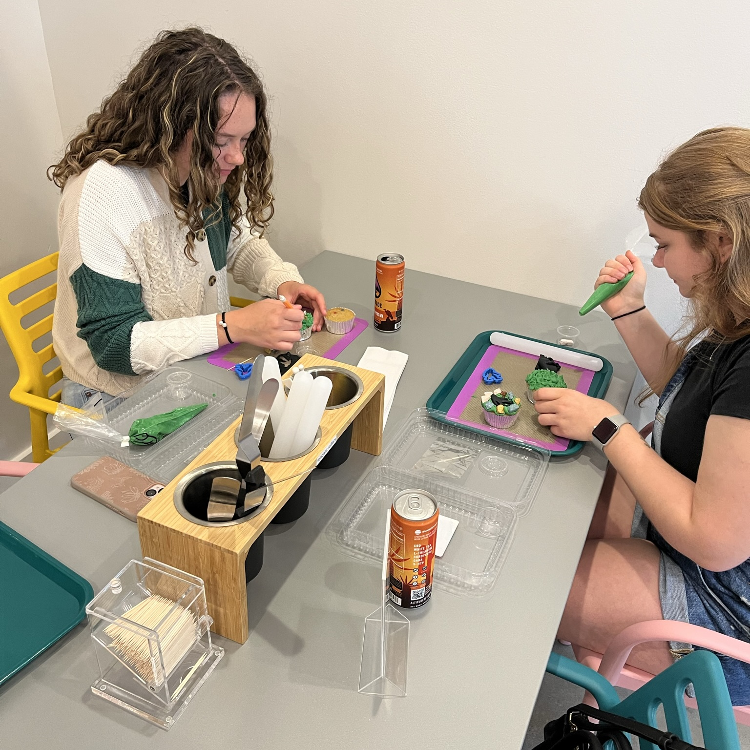 two young ladies decorate cupcakes with frosting and tools, while sipping a canned beverage
