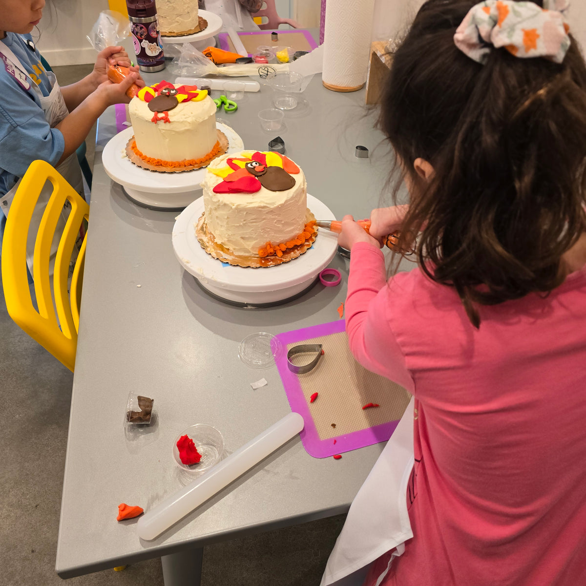 Kids at a table are piping orange frosting onto their white cakes with a fondant turkey on the top.