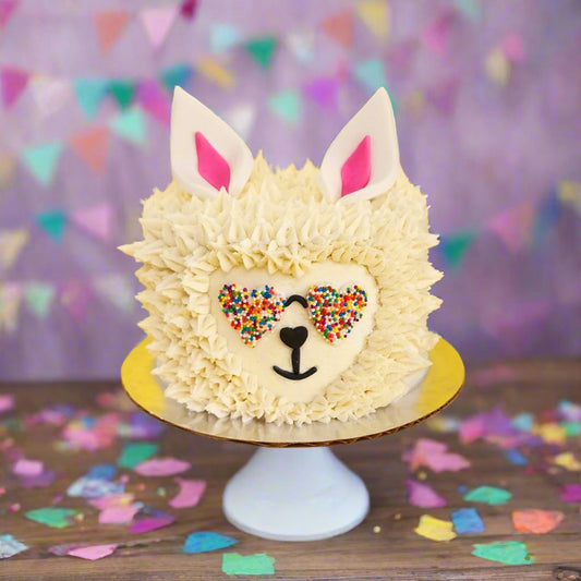 Cute llama cake with white frosting fur and heart-shaped rainbow-sprinkled sunglasses, ready to serve or decorate during open studio time or via a to-go cake kit at Cake Hoopla in Tigard, near Portland.