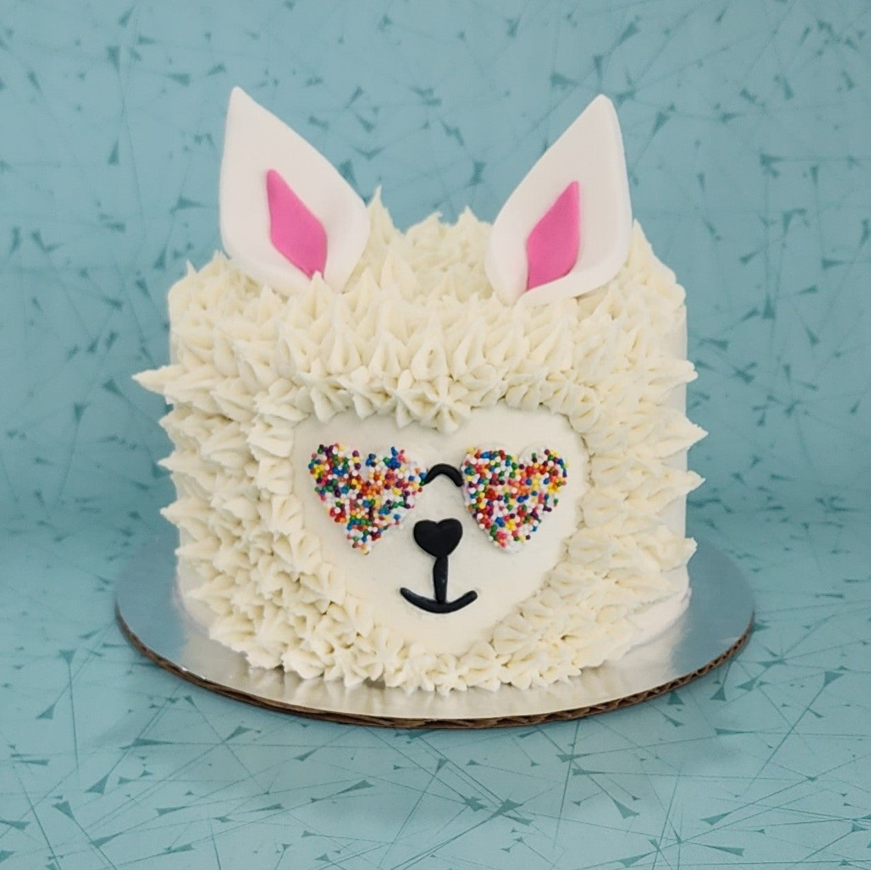 Cute llama cake with white frosting fur, heart-shaped rainbow-sprinkled sunglasses, and white ears with pink centers.