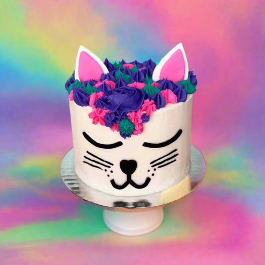 A white cake decorated like a cat face with purple, pink, and teal frosting available as a cake kit or ready-to-serve cake at Cake Hoopla in Tigard, Oregon near Portland.