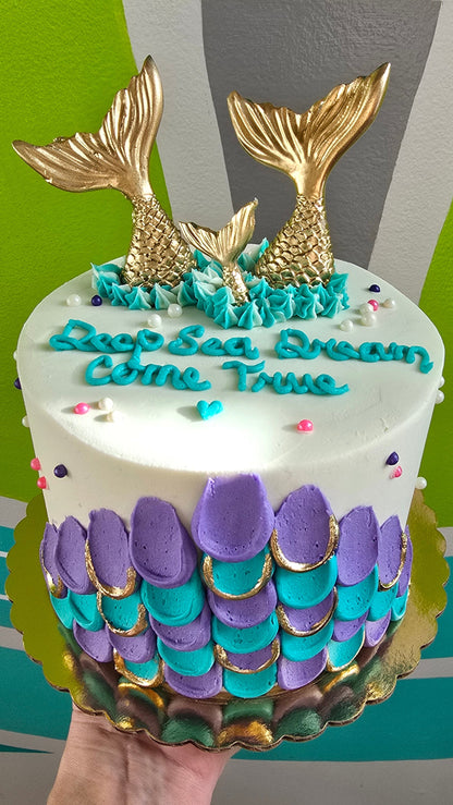 A hand holds a decorated round cake with two gold mermaid tails, purple and teal scalloped scale-like frosting details, and blue icing text reading 'Deep Sea Dreams Come True' against a bright background.