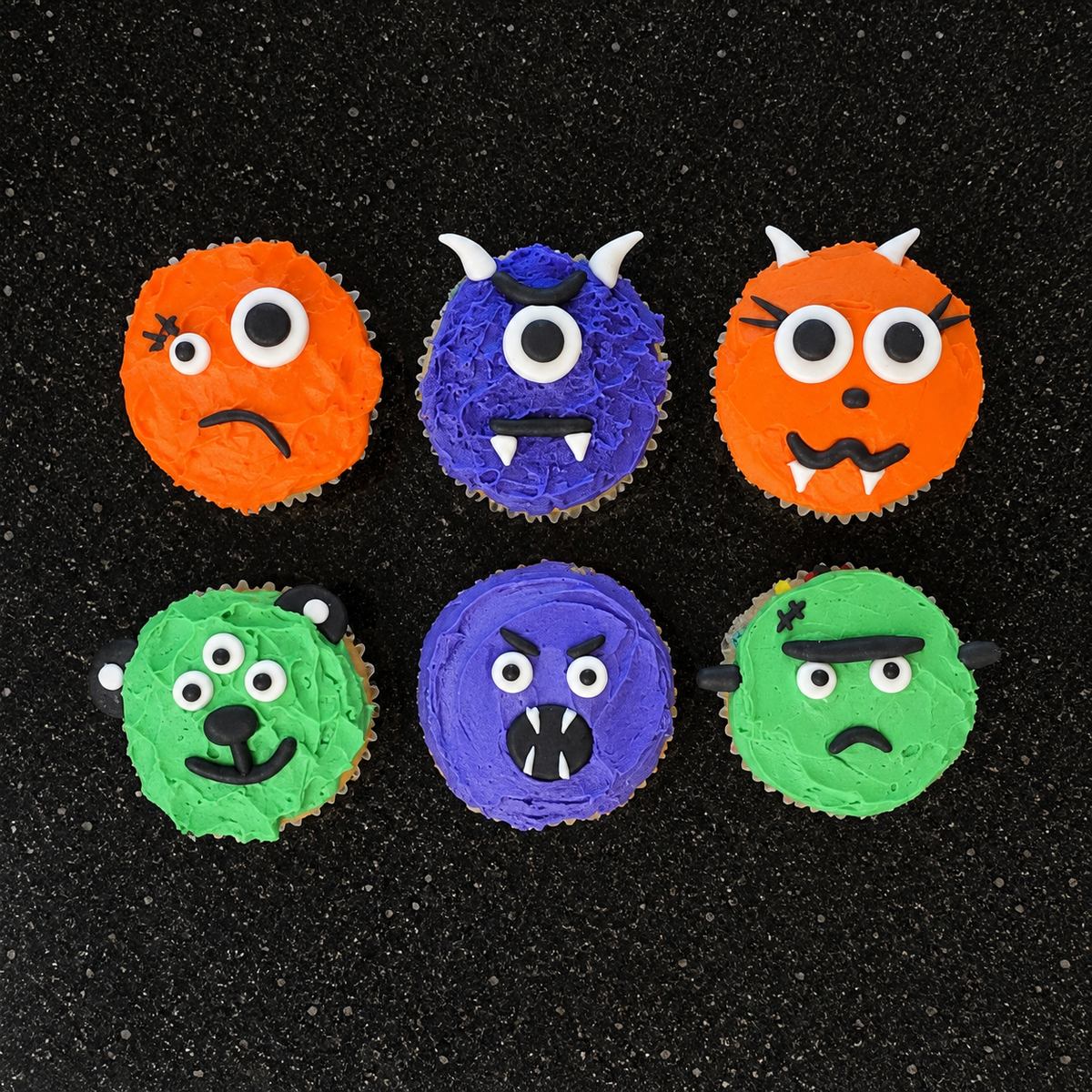 Orange, purple, and green monsters with different scary facial features, like scars, three yes, horns, and fangs, decorated in a Halloween motif on a black stone background.