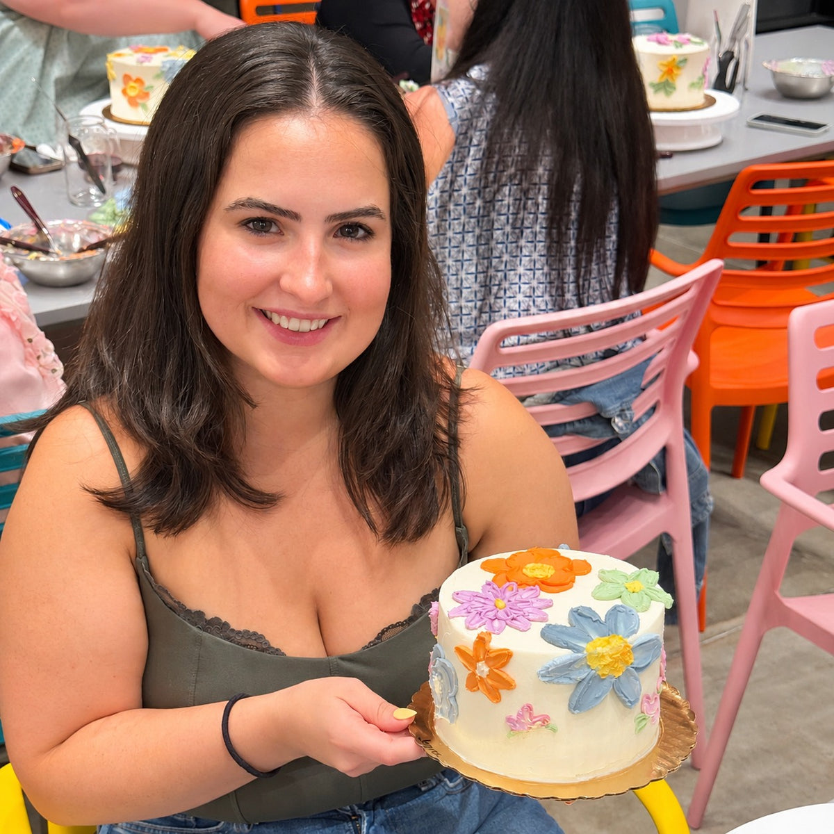 A woman holds a cake frosted white with big blue, pink, orange, and green flowers painted on.