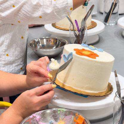 A person tips a white cake to apply blue frosting flowers with a palette knife in a studio setting.