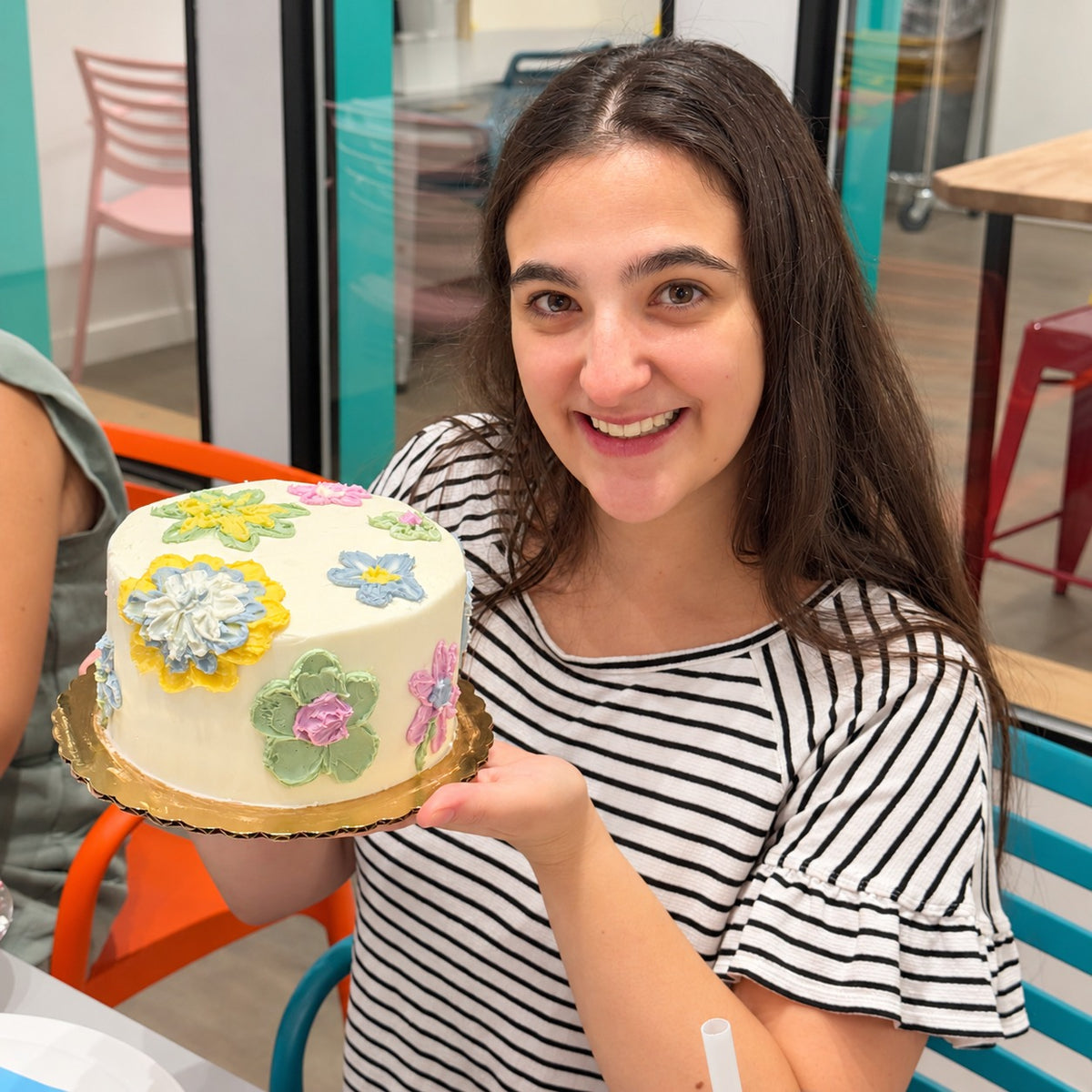 A smiling woman with long dark hair holds up a small round cake decorated with colorful buttercream palette flowers on a white frosting base. 