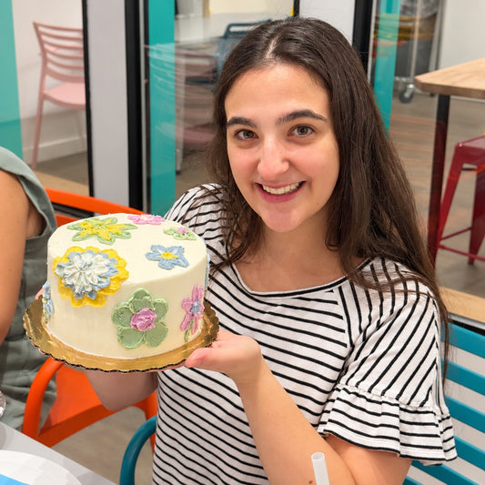 A smiling woman with long dark hair holds up a small round cake decorated with colorful buttercream palette flowers on a white frosting base. 