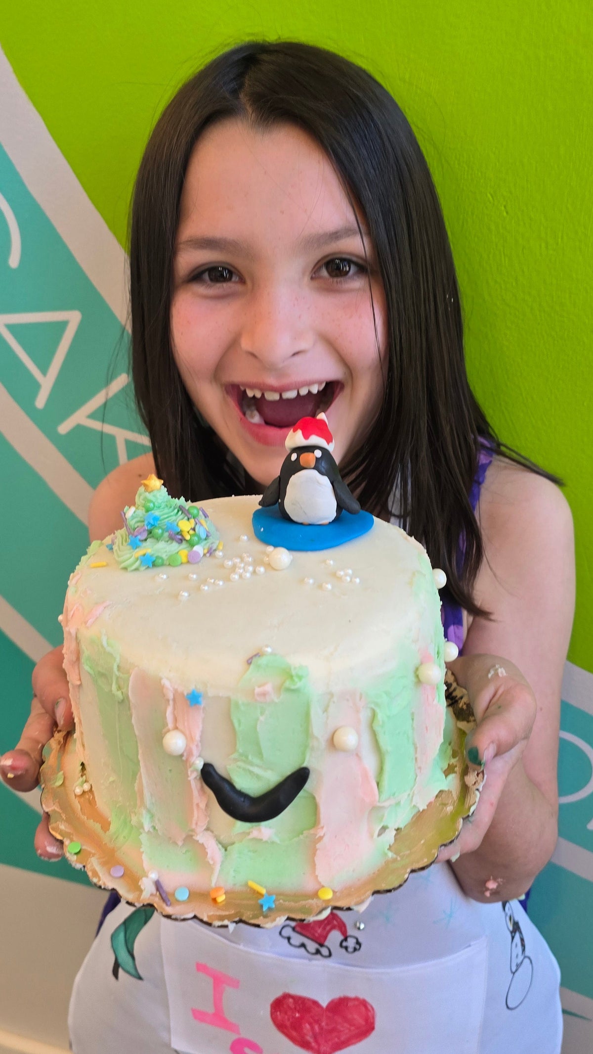 A girl holds a cake striped pastel green and pink topped with a fondant penguin and frosting Christmas tree.