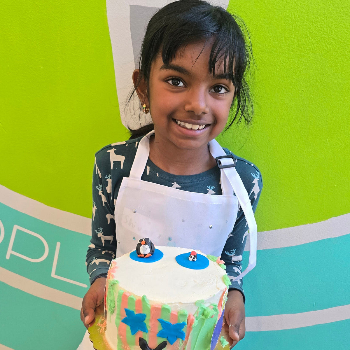 A girl holds up her cake with a penguin on top and a smiling face on on the sides, which are striped with green and red.