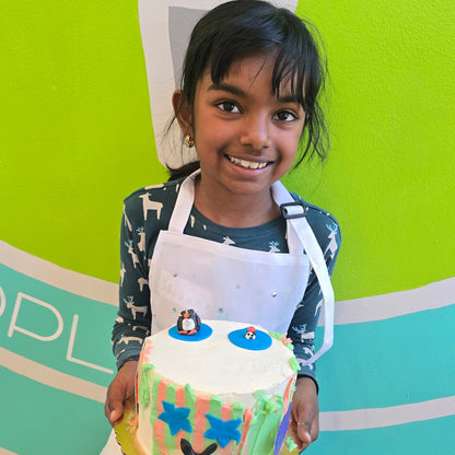 A girl holds up her cake with a penguin on top and a smiling face on on the sides, which are striped with green and red.