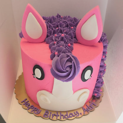 A pink and purple pony-themed birthday cake with white accents, decorated with a purple rosette and pink ears, sitting in a light-colored box. The cake is written 'Happy Birthday, Adri' on the cardboard base.