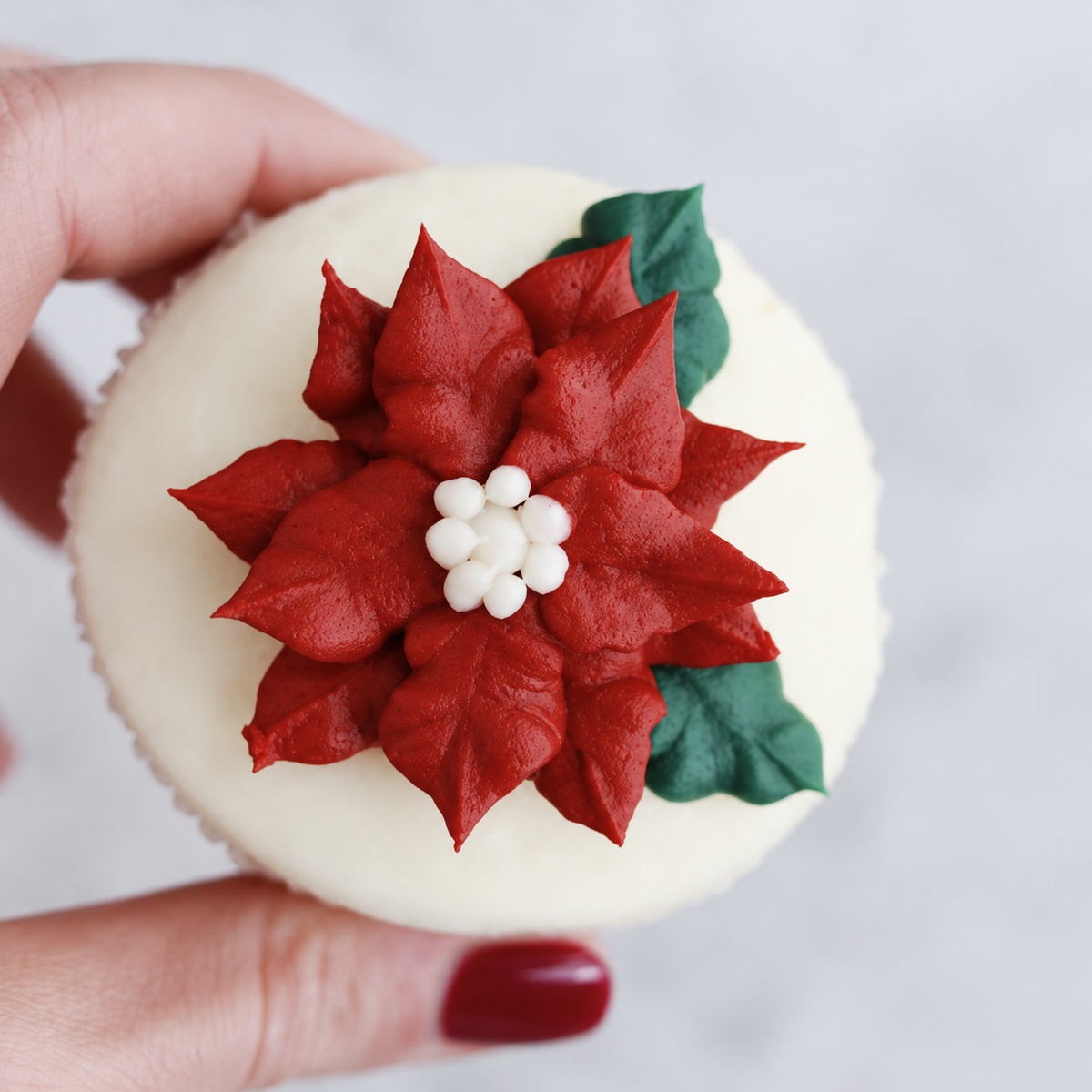 Red poinsettia frosting flower and green leaves across a white-frosted cupcake. 