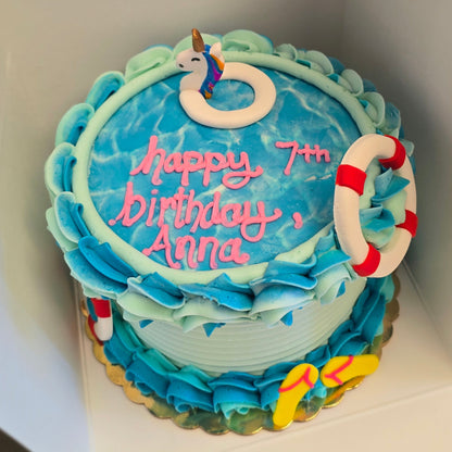 A round pool-themed birthday cake decorated with blue and turquoise ruffled frosting, a white lifebuoy topper, and pink icing text reading 'Happy 7th birthday Anna', shown inside a white box.